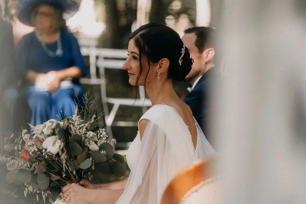 Une mariée très émue lors de l'échange de ses vœux et des alliances. Son maquillage est sophistiqué et élégant, et elle porte un chignon bas, entouré d'une broche à cheveux.